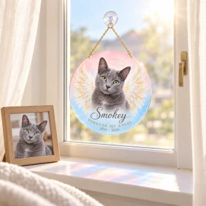 Angel Wings Cat Portrait Suncatcher styled in a lifestyle scene – hanging from a bedroom window latch with sheer white curtains, golden late-afternoon sun glowing through the wings, a framed photo on the dresser below