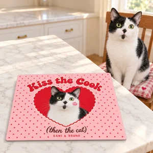 Kiss the Cook Then the Cat Valentine Cutting Board styled in a lifestyle scene – a cheerful Valentine's breakfast nook with a pink striped tea towel, two mugs and a small jar of red roses (well away from the cat), soft morning light