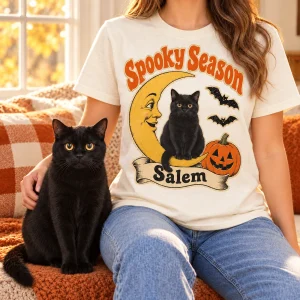 Vintage Die-Cut Halloween Black Cat T-Shirt styled in a lifestyle scene – a person wearing the black tee on a porch step beside carved pumpkins (unlit, battery fairy lights only) and a straw bale, golden-hour autumn light